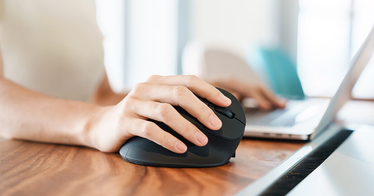 Woman using ergonomic mouse at desk beside laptop.